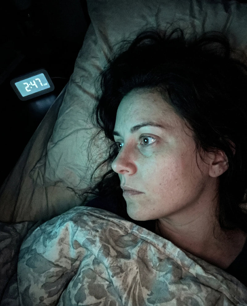A tight three-quarter shot of a woman lying on her side in the dark — not crying, just awake. One eye visible and wide open, expression flat and depleted but unable to close. The only light is a faint blue-green glow from a baby monitor or a face-down phone spilling across the pillow and her cheek; a clock or the monitor reads 2:47. Rumpled, real bedding. Shot on a phone or graded to look phone-shot, slight grain, no gloss.